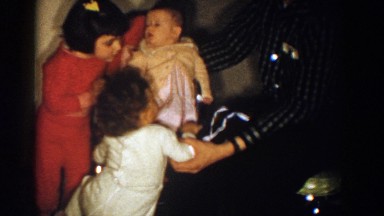 1958: Babysitting in High Chair Against Wall Surrounded by Two Children