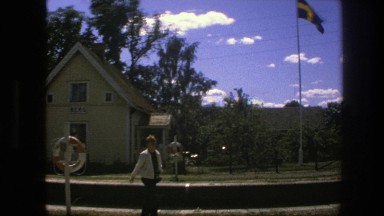 1975: a woman walks by a little yellow house on dirt path with the swedish flag