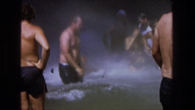 1969: a group of men standing in bathing suits under a waterfall next to rocks