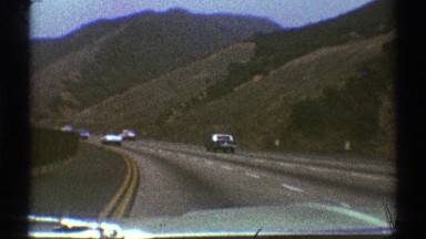 1969: cars riding on a road next a mountain covered in green grass and trees