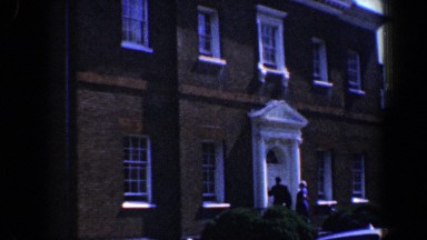 1961: cars line the street in front of a large colonial house where two men wait