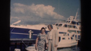 1961: couple having their photo made in front of a large boat on water.