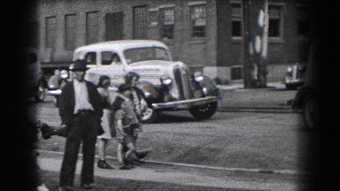 1938: Man and Children Stand Near Curb as Vintage Cars Drive By at Town Intersection