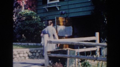 1960: a woman in a dress and pinafore playfully shakes wine bottle at camera