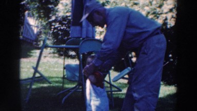 1964: Grandfather Wearing Hat Helping Grandchild Walk Outside Tustin
