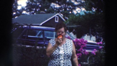 1964: Woman Eating Fruit While Waving to Camera in Front of Flowers Tustin