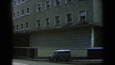 1960: people walking in the street near a tall structure and a vehicle parked