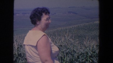 1959: a middle aged woman in a dress and glasses stands in a corn field