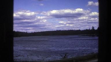 1975: Lake Rimmed With Trees Under Blue Sky Filled With Puffy White Clouds
