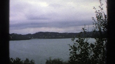 1976: a view of a lake with mountains on the horizon trees and storm clouds