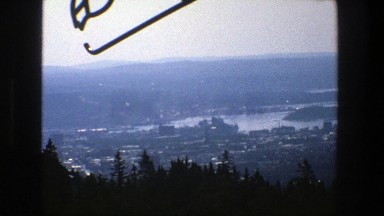 1975: view of a developed city on a river from atop a hill behind pine trees