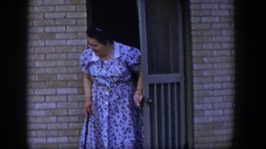 1954: an older woman in a purple dress and cane in hand steps outside her home