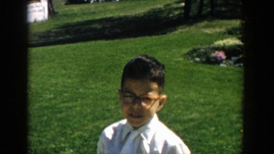 1957: little boy in orange framed glasses walks past family car in front yard