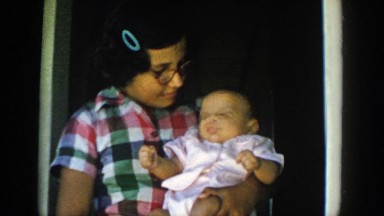 1958: girl carries baby in crook of her arm facing forward and makes baby wave
