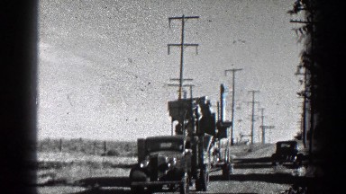 1936: Old Model Truck Pulling Trailer with Machinery Through Village Road