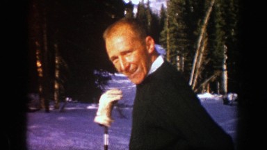 1961: man putting on jacket with ski pole and glove beside him, standing on a