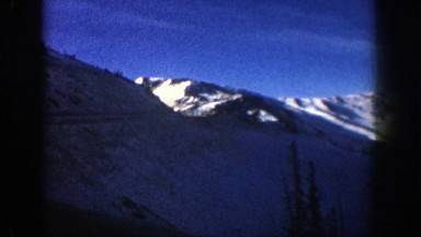 1961: car ascending snow-covered mountain range under blue sky with wispy, long