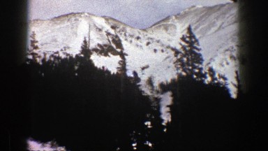 1961: panorama of a snowy mountain valley with trees and jagged peaks and a road