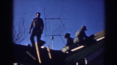 1958: three men on house roof laying shingles below antennae, bare branches and