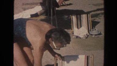 1961: middle aged woman setting up a beach chair at a beach on a sunny day