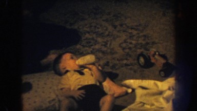 1959: a set of male twins dressed up at home with mom laying down with a bottle.