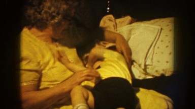 1959: baby laying on the bed and drinking milk from the bottle with mom and dad.