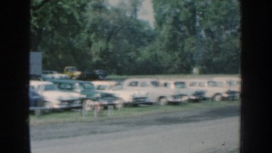 1959: row of cars fill a parking lot between trucks, trees and building for