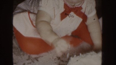 1955: a very young child sits on the floor eating a cake with family around