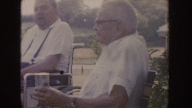 1955: an old man sits in a chair next to another man while drinking from a can.