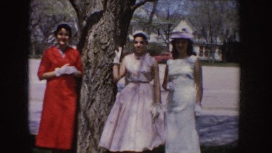 1955: three dressed-up women smile and pose in a row by tree near residential