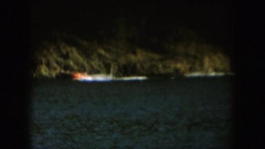 1954: an orange speedboat pulling along two waterskiers close to a rocky shore.