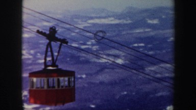 1962: a bright red lift car carries people high above snow capped mountains.
