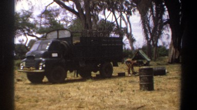 1969: Man in Grassy Area Bending Behind Big Black Truck Looking for Something