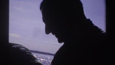 1969: dimly lit people move in front of a brightly lit seascape, one turning to