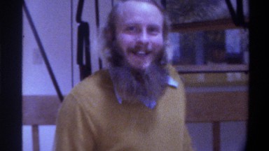 1975: a smiling man with a beard sits at a dinner table by himself while smoking