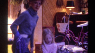 1975: girl tears wrapping paper off rolling box on floor and is helped by