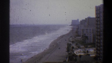 1973: low populated beach, with man walking along the shore, and buildings NEW