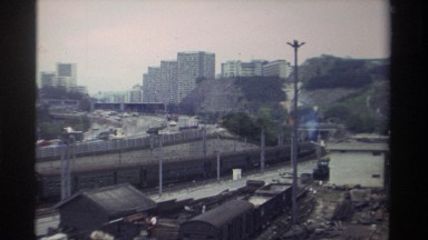 1982: Vehicles and Train Curve on Road and Tracks Between Rail Yard and City