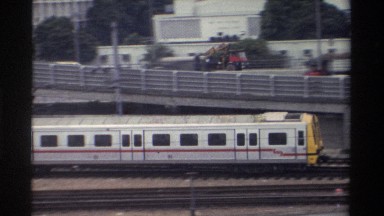1982: silver train rolling along tracks at modest speed with cars driving in