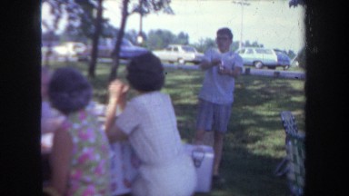 1968: large family picnic in a park, car driving across in the background