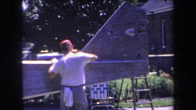 1969: Man Wearing White T-Shirt Polishing Large Metal Piece in Backyard