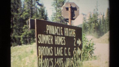 1980: sign posted along road lists locations with arrows in a forest. WESTERN