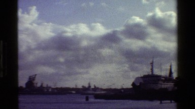 1983: a camera shot spans and shows several ships at the harbor, a blue sea, and