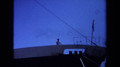 1971: a man standing on top of a large navy ship while people walk pass. MAINE