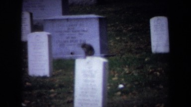 1970: dark squirrel rests and scratches with hind leg on top of headstone