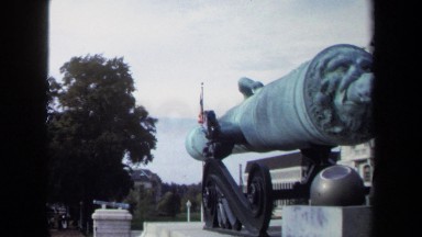 1970: People Walk on Wide Stone Path Between Elevated Cannons and Flags on Poles