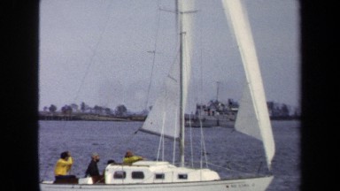 1970: white sailboat with passengers in back passes gray ship with shore