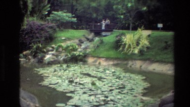 1984: Two People Stand on Wooden Bridge Over Stream in Lush Garden with Pond