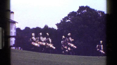 1983: tribesmen running up a hill with ornate drums past a building KENYA