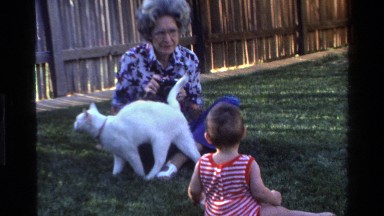 1977: an old lady and a baby sit in a yard playing with a white cat CALIFORNIA